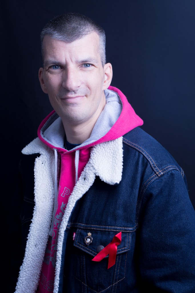 Photo of HIVBlogger with a pink hoodie and a blue denim jacket with a red ribbon on. 