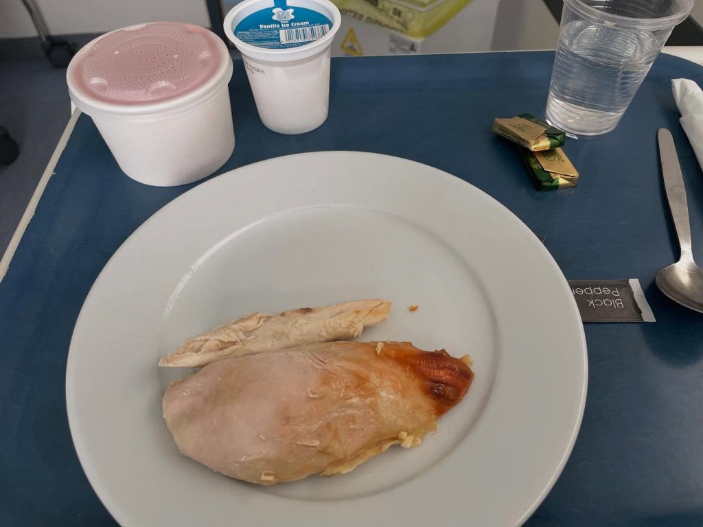 A tray with a white plate with chicken breast on it, with a glass of water, a spoon, two butter packs, and two plastic containers of jelly and ice cream. 