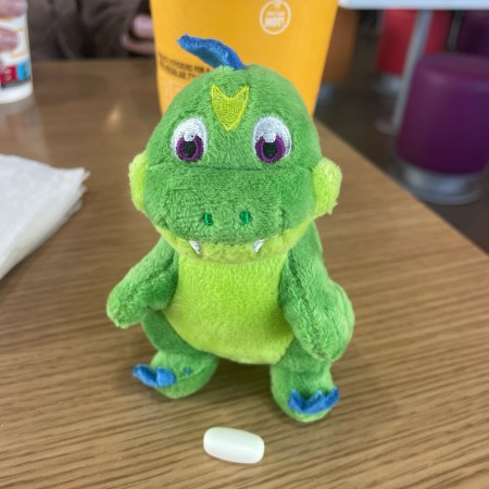 A green dragon plushie with white tablet sitting in front of him on a table.